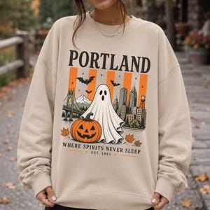 Könnte beinhalten: Ein beigefarbenes Sweatshirt mit Rundhalsausschnitt und einem Grafikdruck mit dem Text PORTLAND, WHERE SPIRITS NEVER SLEEP, EST. 1851. Das Design zeigt ein Gespenst, einen Kürbis, Fledermäuse, Herbstblätter, eine Brücke und eine Skyline.