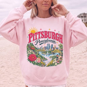May include: A light pink crewneck sweatshirt featuring a colourful graphic print of the Pittsburgh, Pennsylvania skyline. The design includes a yellow sun, red text, a red funicular, yellow bridges, a river, and vibrant floral accents.