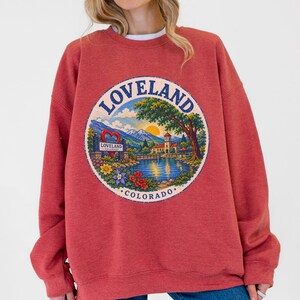 Könnte beinhalten: Ein ziegelrotes Sweatshirt mit Rundhalsausschnitt und einem kreisförmigen Aufdruck auf der Brust. Das Motiv zeigt eine Landschaft mit Bergen, einem See, einem Gebäude und einem großen Baum sowie den Text LOVELAND COLORADO und ein Herzsymbol.