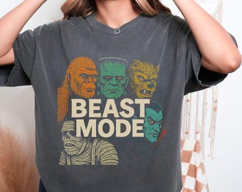 Pode incluir: T-shirt cinza escuro com um gráfico de rostos de monstros e o texto "BEAST MODE". O design inclui um Sasquatch, o monstro de Frankenstein, um lobisomem, uma múmia e um vampiro. A camisa tem gola redonda clássica e mangas curtas.
