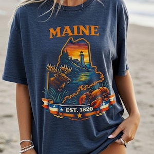 Puede incluir: Camiseta azul marino con un gráfico del estado de Maine. El diseño incluye un faro, un alce y una langosta, con la palabra "MAINE" en naranja y "EST. 1820" en una pancarta. La camiseta es de corte holgado.