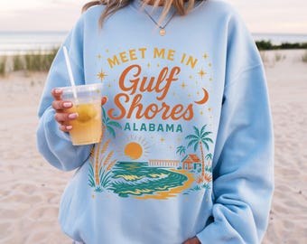 May include: Light blue sweatshirt with the text "MEET ME IN Gulf Shores ALABAMA" in orange and yellow. The graphic includes a beach scene with palm trees, a pier, and a sunset. The person is holding a clear plastic cup with a straw and a yellow beverage.