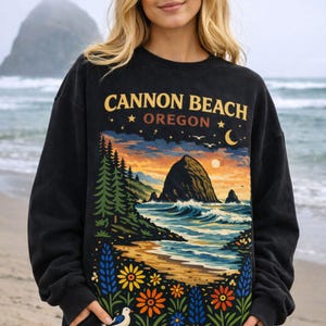 May include: Black sweatshirt with a colorful graphic of Cannon Beach, Oregon. The design features a coastal landscape with a large rock formation, ocean waves, and a sunset sky. The text "CANNON BEACH OREGON" is printed across the top.
