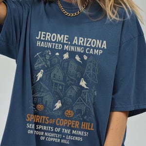 Op de afbeelding: Marineblauw T-shirt met een grafisch ontwerp met de tekst "JEROME, ARIZONA HAUNTED MINING CAMP". Het ontwerp bevat illustraties van gebouwen, geesten en pompoenen. De tekst "SPIRITS OF COPPER HILL" is ook zichtbaar.