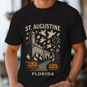 Può includere: T-shirt nera con grafica a tema Halloween. Il design include il testo "ST. AUGUSTINE FLORIDA" sopra e sotto una raffigurazione di un forte, fantasmi e zucche. Sono inclusi anche una palma e un lampione.