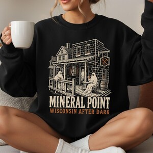 May include: A black crewneck sweatshirt featuring a graphic illustration of a building with ghosts on a porch, a lantern, and a barrel. The text reads "MINERAL POINT" and "WISCONSIN AFTER DARK" in a vintage-style font with a pickaxe sign.