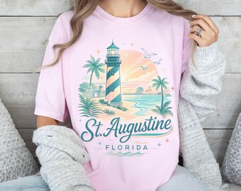 May include: A light pink t-shirt featuring a coastal design with a lighthouse, palm trees, and the text "St. Augustine Florida". The lighthouse has blue and white stripes. The design is set against a sunset backdrop.