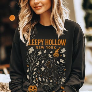 May include: Black sweatshirt with the text "Sleepy Hollow New York" in orange. The design features a headless horseman, ghosts, a jack-o'-lantern, and a tree. A spooky Halloween-themed graphic.