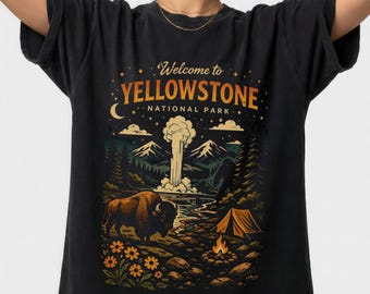 May include: Black t-shirt featuring a colorful graphic of Yellowstone National Park. The design includes a bison, mountains, a geyser, a campfire, and the text "Welcome to Yellowstone National Park."