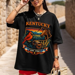 Może przedstawiać: Czarna koszulka z grafiką Kentucky. Projekt przedstawia czerwonego kardynała, konia, beczkę oraz napisy "KENTUCKY" i "EST. 1792". Grafika jest osadzona na tle krajobrazu z zachodem słońca.