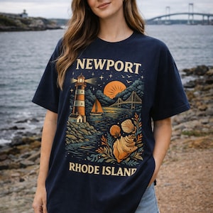 以下が含まれることがあります： ニューポート、ロードアイランドのデザインが施された濃いネイビーブルーのTシャツ。灯台、ヨット、橋、貝殻が夕焼けと星空を背景に描かれています。「NEWPORT」と「RHODE ISLAND」の文字が大きく表示されています。