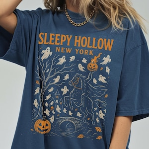 May include: A navy blue t-shirt with an orange graphic design featuring the text "Sleepy Hollow New York". The design includes a headless horseman, ghosts, a pumpkin, a bridge, and a tree.