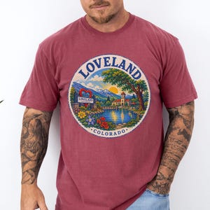 May include: A maroon short-sleeve t-shirt featuring a circular graphic print of a landscape in Loveland, Colorado. The design includes a red heart, a lake, mountains, trees, flowers, and a building with the text LOVELAND COLORADO.