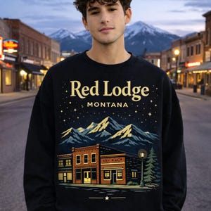 May include: Black sweatshirt with a graphic of a town scene with the text "Red Lodge MONTANA". The design features a night sky, mountains, and a building with a street lamp and a pine tree.