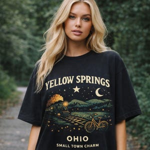 May include: Black t-shirt with the text "YELLOW SPRINGS" above a graphic of a train, bicycle, and night sky. The text "OHIO SMALL TOWN CHARM" is below the graphic. The shirt is worn by a person with blonde hair.