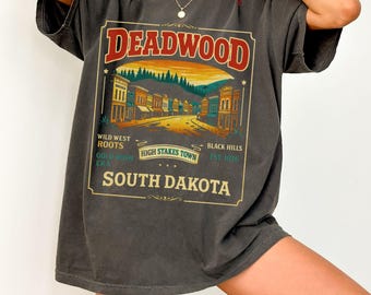 May include: Dark gray t-shirt featuring a vintage-style graphic with the word "DEADWOOD" in red, and a landscape scene. The graphic also includes text that reads "Wild West Roots", "High Stakes Town", and "South Dakota".