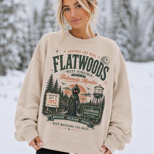 May include: A beige crewneck sweatshirt featuring a graphic print with the text "FLATWOODS WEST VIRGINIA" and "LEGENDS ARE REAL". The design includes a hooded figure with glowing eyes, a water tower, pine trees, and the text "SIGHTED SEPT. 12 1952".