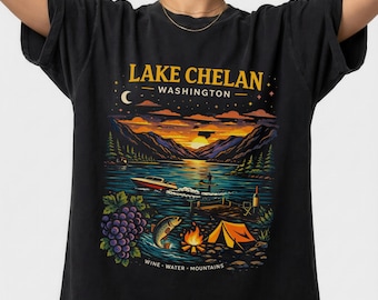 May include: Black t-shirt featuring a colorful graphic of Lake Chelan, Washington. The design includes a lake scene with a boat, mountains, a campfire, and the text "Wine - Water - Mountains". A crescent moon and stars are also visible.