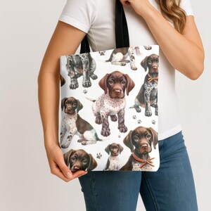 May include: White tote bag featuring a repeating pattern of watercolor-style German Shorthaired Pointer puppies in various poses. The puppies are brown, white, and black with paw prints scattered across the white background. The bag has black handles.