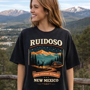 Könnte beinhalten: Schwarzes T-Shirt mit einem Berglandschaftsdesign, einem Hirsch und einer Hütte. Der Text "RUIDOSO" steht oben, darunter "HIGH COUNTRY RETREAT" und "NEW MEXICO". Das T-Shirt wird von einer Person mit langen blonden Haaren getragen.