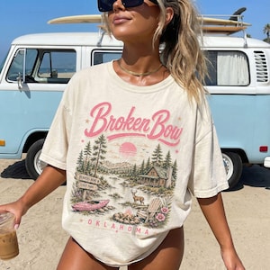May include: A cream-colored short-sleeve t-shirt featuring a graphic print with the text Broken Bow and Oklahoma. The design depicts a forest landscape with a cabin, a lake, a kayak, a deer, a campfire, and a sign reading Beavers Bend State Park.