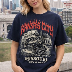 Op de afbeelding: Marineblauwe t-shirt met een vintage illustratie van een stoomtrein en de tekst "KANSAS CITY" in rode vlammen. Daaronder staat "MISSOURI" en de slogan "SHOW ME STATE". De t-shirt heeft een ronde hals.