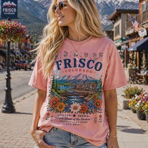 Pode incluir: Uma camiseta rosa empoeirada com um gráfico de Frisco, Colorado, apresentando uma cena de montanha, rio e borda floral. O texto "FRISCO COLORADO" está acima da imagem, com "Main Street of the Rockies" e "EST. 1879" abaixo.