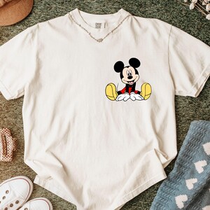 May include: Off-white t-shirt featuring a seated Mickey Mouse graphic. The cartoon character is depicted with black ears, a red jacket, yellow shoes, and white gloves. The shirt is made of a soft material and has a V-neck collar.