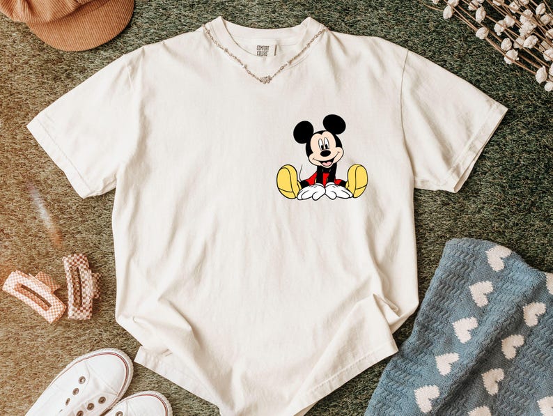May include: Off-white t-shirt featuring a seated Mickey Mouse graphic. The cartoon character is depicted with black ears, a red jacket, yellow shoes, and white gloves. The shirt is made of a soft material and has a V-neck collar.