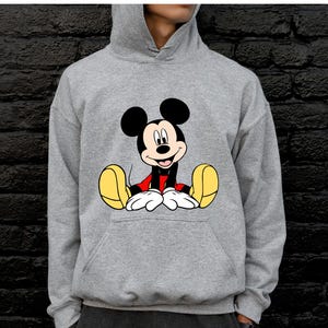 May include: A gray hooded sweatshirt featuring a large graphic of Mickey Mouse. The cartoon character is depicted in his classic pose, with black ears, a red shirt, white gloves, and yellow shoes. The sweatshirt has a front pocket and a drawstring hood.