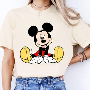 May include: Cream-colored t-shirt featuring a smiling Mickey Mouse graphic. The cartoon character is depicted with black ears, a red shirt, yellow shoes, and white gloves. The shirt is made of a soft material.