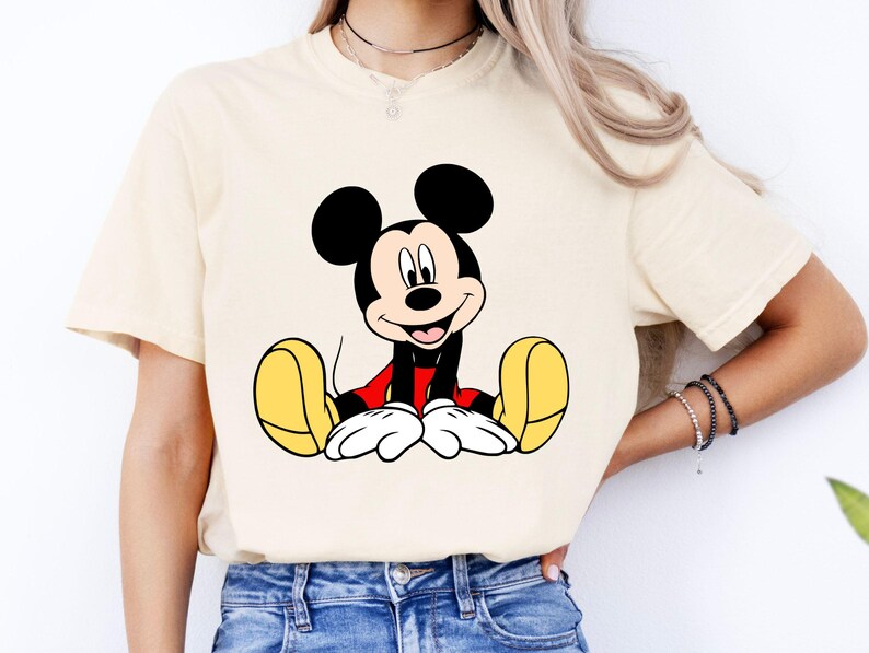 May include: Cream-colored t-shirt featuring a smiling Mickey Mouse graphic. The cartoon character is depicted with black ears, a red shirt, yellow shoes, and white gloves. The shirt is made of a soft material.