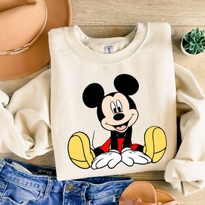 May include: Cream-colored sweatshirt featuring a smiling Mickey Mouse graphic. Mickey is depicted with black ears, a red shirt, yellow shoes, and white gloves. The sweatshirt is paired with blue denim shorts, a tan hat, and sandals.