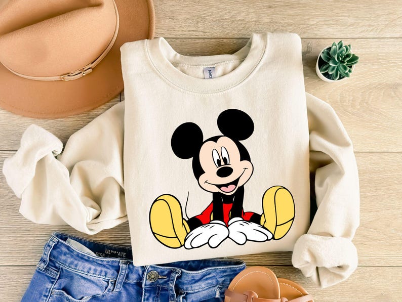 May include: Cream-colored sweatshirt featuring a smiling Mickey Mouse graphic. Mickey is depicted with black ears, a red shirt, yellow shoes, and white gloves. The sweatshirt is paired with blue denim shorts, a tan hat, and sandals.