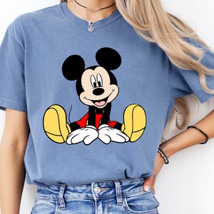 May include: A blue t-shirt featuring a graphic of Mickey Mouse. The cartoon character is depicted with black ears, a red shirt, white gloves, and yellow shoes. The shirt is a solid blue color.
