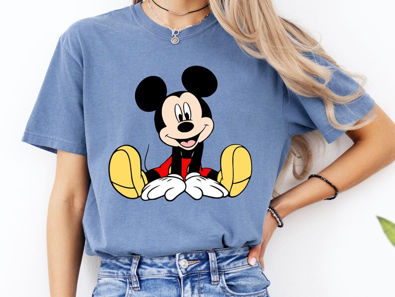 May include: A blue t-shirt featuring a graphic of Mickey Mouse. The cartoon character is depicted with black ears, a red shirt, white gloves, and yellow shoes. The shirt is a solid blue color.