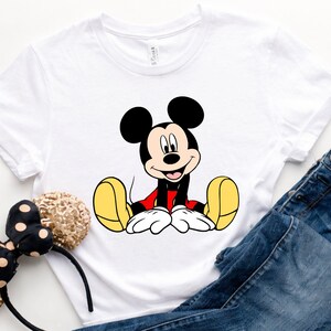 May include: White t-shirt featuring a seated Mickey Mouse graphic in black, red, yellow, and white. The shirt is paired with blue denim jeans and a black and gold polka dot headband.