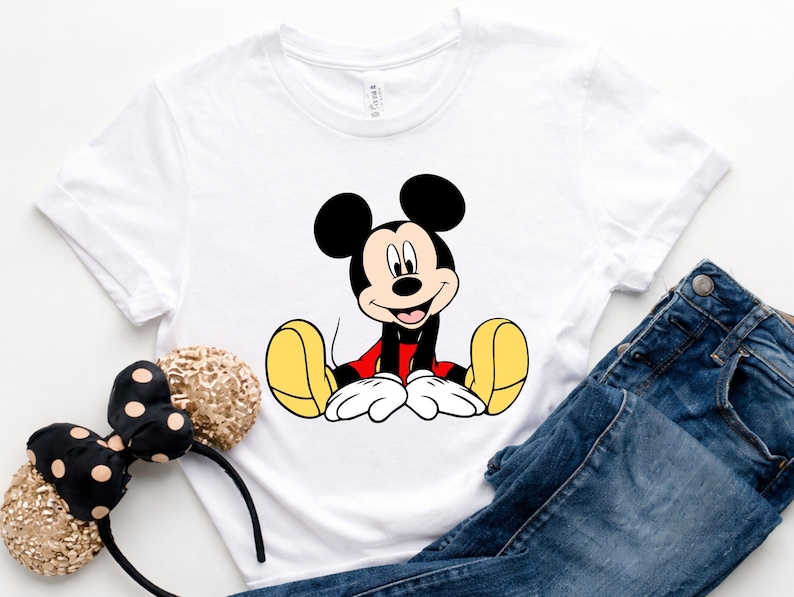 May include: White t-shirt featuring a seated Mickey Mouse graphic in black, red, yellow, and white. The shirt is paired with blue denim jeans and a black and gold polka dot headband.