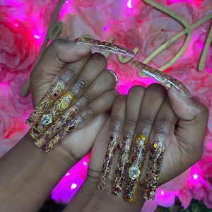 Puede incluir: Uñas acrílicas largas y transparentes con purpurina dorada y flores secas incrustadas en el esmalte de uñas. Las uñas están decoradas con pequeños diamantes de imitación.
