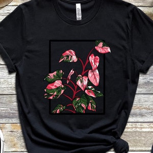 以下が含まれることがあります： 黒いTシャツに、ピンクと緑の植物が黒い枠の中に描かれたグラフィックプリントが施されています。