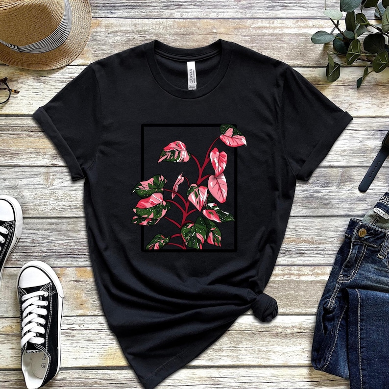 以下が含まれることがあります： 黒いTシャツに、ピンクと緑の植物が黒い枠の中に描かれたグラフィックプリントが施されています。