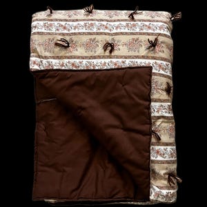 May include: A rectangular quilt featuring a beige background with horizontal stripes of floral patterns in shades of brown and orange. The quilt is accented with small dark brown yarn tassels and has a solid dark brown underside folded over.