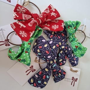 May include: A collection of four fabric hair ties featuring festive holiday patterns. One bow is red with gingerbread men and snowflakes, one is green with holly leaves and berries, and two are dark blue with colourful Christmas light bulb designs.