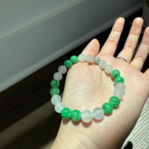 May include: A white and green jade bead bracelet with a silver charm. The bracelet is made of round beads and has a simple design.
