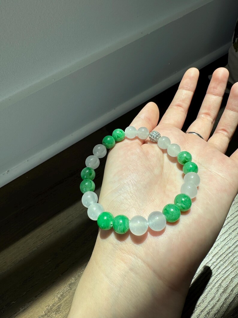 May include: A white and green jade bead bracelet with a silver charm. The bracelet is made of round beads and has a simple design.