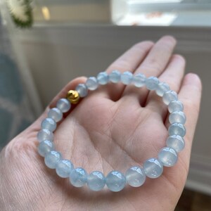 K&ouml;nnte beinhalten: Ein hellblaues Edelstein-Armband mit einem goldenen Perlenakzent. Das Armband besteht aus runden Perlen und wird am Handgelenk getragen.