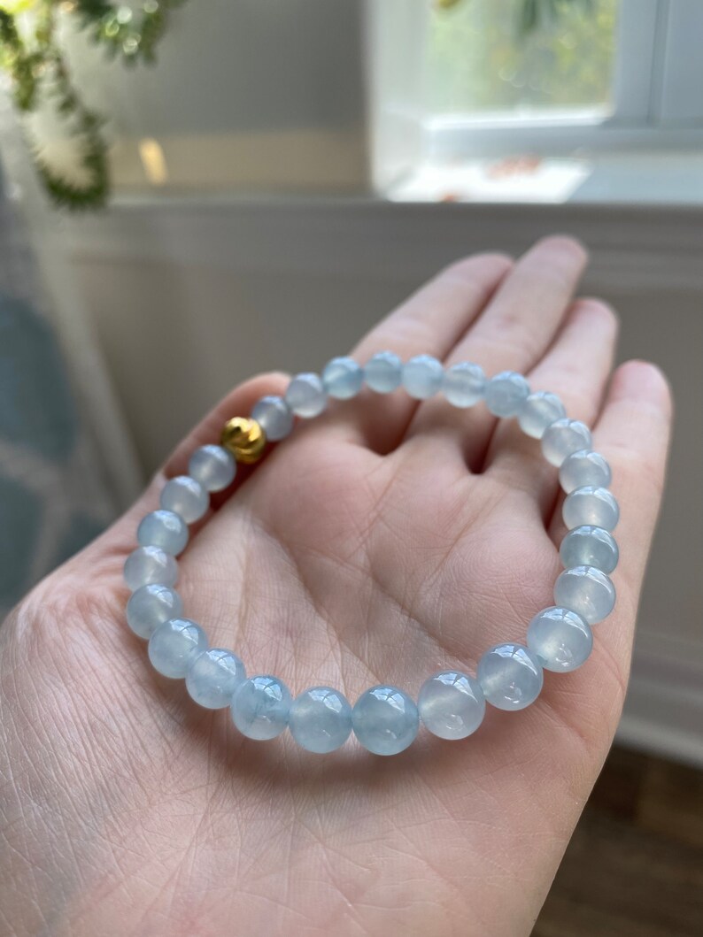 K&ouml;nnte beinhalten: Ein hellblaues Edelstein-Armband mit einem goldenen Perlenakzent. Das Armband besteht aus runden Perlen und wird am Handgelenk getragen.
