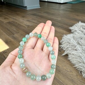 Pode incluir: Um bracelete de contas de jade verde com um detalhe de pedra preciosa dourada e vermelha. O bracelete &eacute; feito de contas redondas e &eacute; usado no pulso.