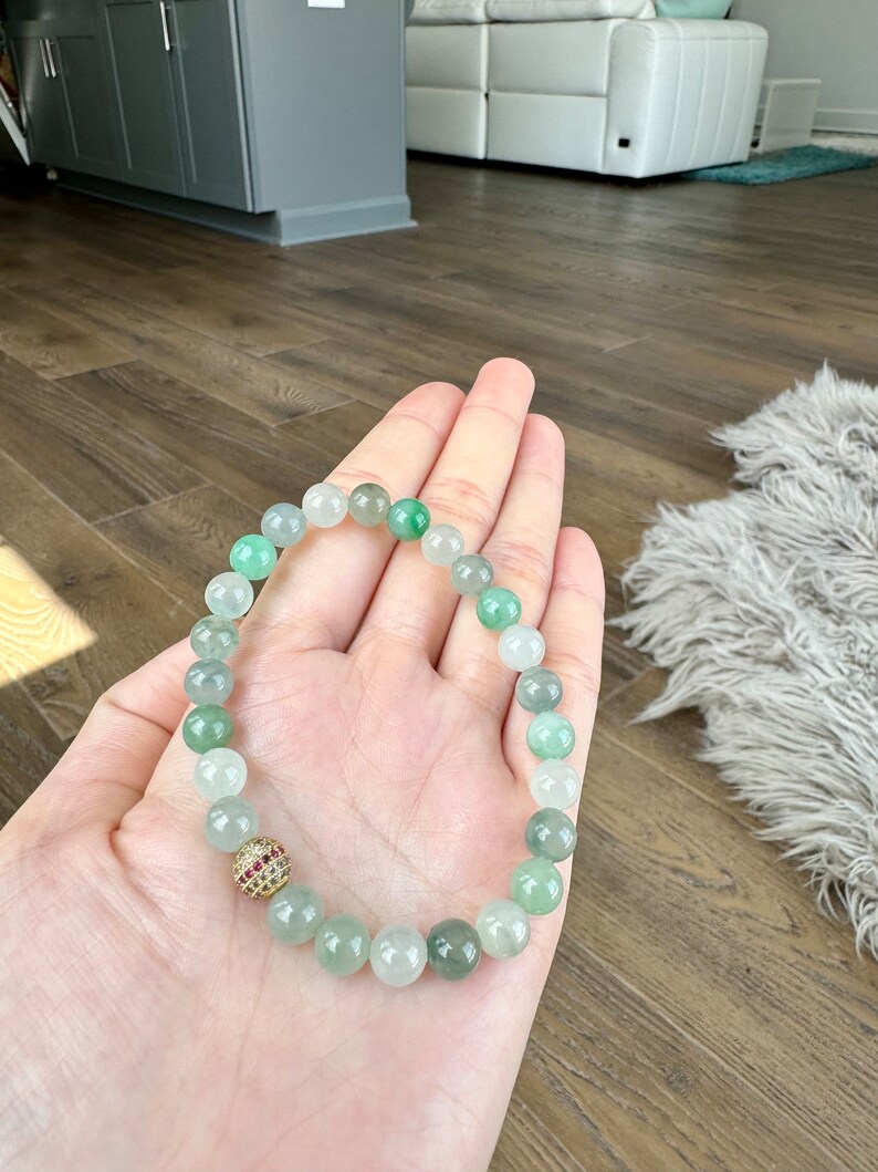 Pode incluir: Um bracelete de contas de jade verde com um detalhe de pedra preciosa dourada e vermelha. O bracelete &eacute; feito de contas redondas e &eacute; usado no pulso.