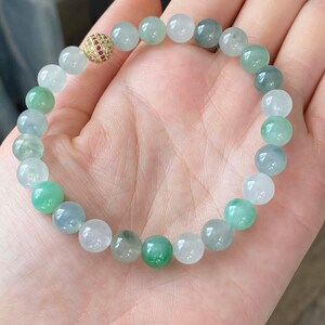 Pode incluir: Um bracelete de jade&iacute;ta com uma conta de ouro e pedra vermelha. O bracelete &eacute; feito de contas redondas em tons de verde e branco.